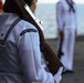 A U.S. Navy sailor in dress white uniform is shown from behind, wearing a shoulder patch reading 'USS DWIGHT D. EISENHOWER'. The sailor appears to be participating in a formal military ceremony or parade, holding a rifle, with other sailors visible in the background near what appears to be a body of water. The setting suggests an outdoor naval ceremony, likely on a pier or flight deck.