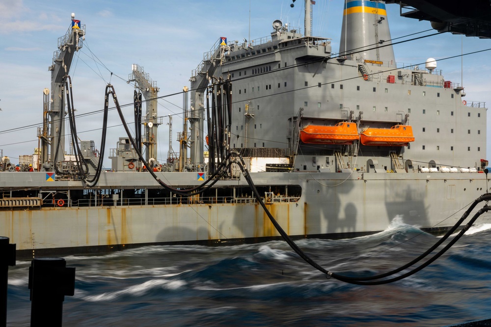 The image shows a replenishment-at-sea (RAS) operation between two naval vessels underway on open ocean. Large black fuel/transfer hoses are visibly connected between the ships, suspended in arcing configurations typical of underway replenishment. The supply vessel, painted gray with orange lifeboats and a blue-and-yellow funnel marking, appears to be a Military Sealift Command (MSC) replenishment oiler.