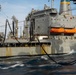 The image shows a replenishment-at-sea (RAS) operation between two naval vessels underway on open ocean. Large black fuel/transfer hoses are visibly connected between the ships, suspended in arcing configurations typical of underway replenishment. The supply vessel, painted gray with orange lifeboats and a blue-and-yellow funnel marking, appears to be a Military Sealift Command (MSC) replenishment oiler.