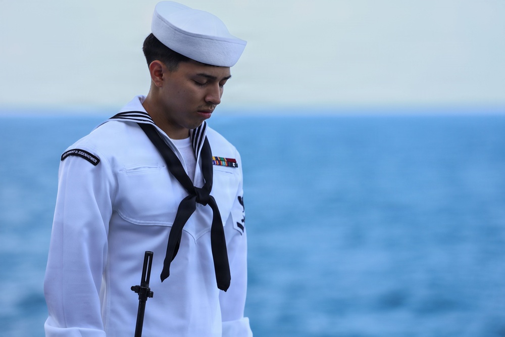 A U.S. Navy sailor in dress white uniform stands at attention with head bowed, holding a rifle, against an ocean backdrop. The shoulder patch reads 'USS Dwight D. Eisenhower,' indicating assignment to the aircraft carrier CVN-69. The solemn posture and formal attire suggest participation in a ceremonial event, possibly a burial at sea or memorial service.