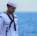 A U.S. Navy sailor in dress white uniform stands at attention with head bowed, holding a rifle, against an ocean backdrop. The shoulder patch reads 'USS Dwight D. Eisenhower,' indicating assignment to the aircraft carrier CVN-69. The solemn posture and formal attire suggest participation in a ceremonial event, possibly a burial at sea or memorial service.