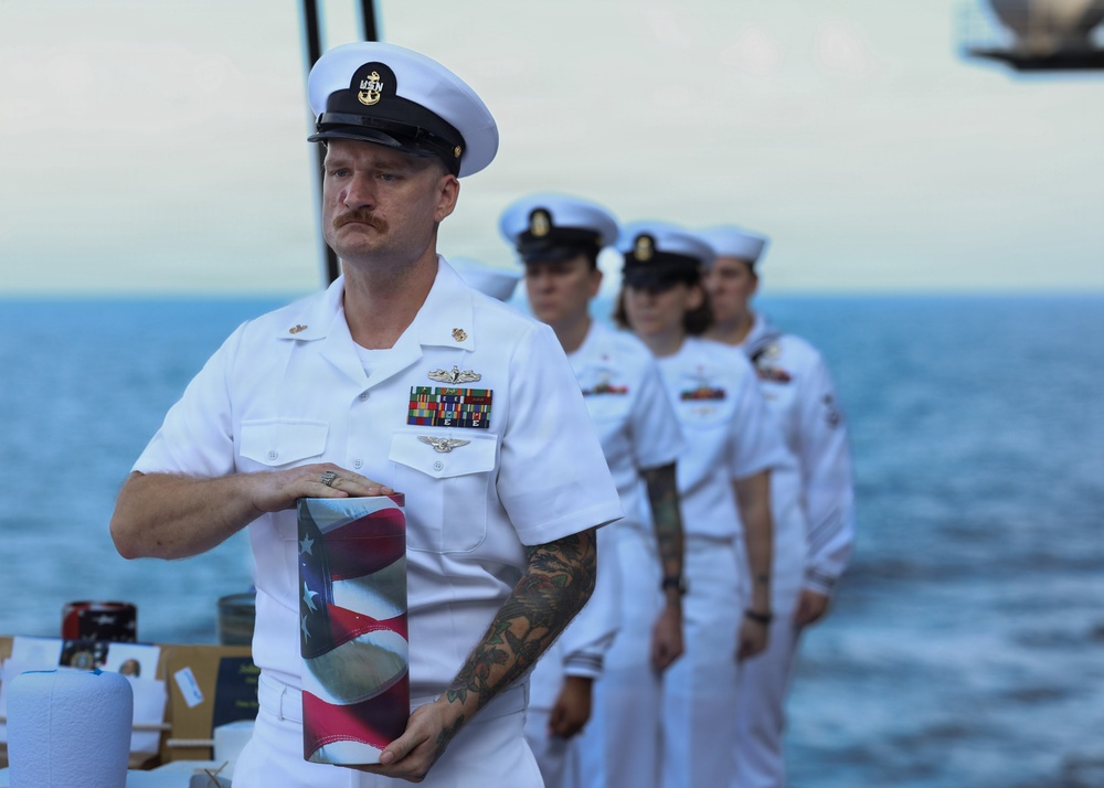 A U.S. Navy senior enlisted sailor in dress white uniform carries a folded American flag during what appears to be a formal military burial at sea ceremony. He is leading a line of fellow sailors also in dress whites aboard a naval vessel with open ocean visible in the background. The solemn expressions and ceremonial flag suggest this is a funeral or memorial service.
