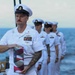 A U.S. Navy senior enlisted sailor in dress white uniform carries a folded American flag during what appears to be a formal military burial at sea ceremony. He is leading a line of fellow sailors also in dress whites aboard a naval vessel with open ocean visible in the background. The solemn expressions and ceremonial flag suggest this is a funeral or memorial service.