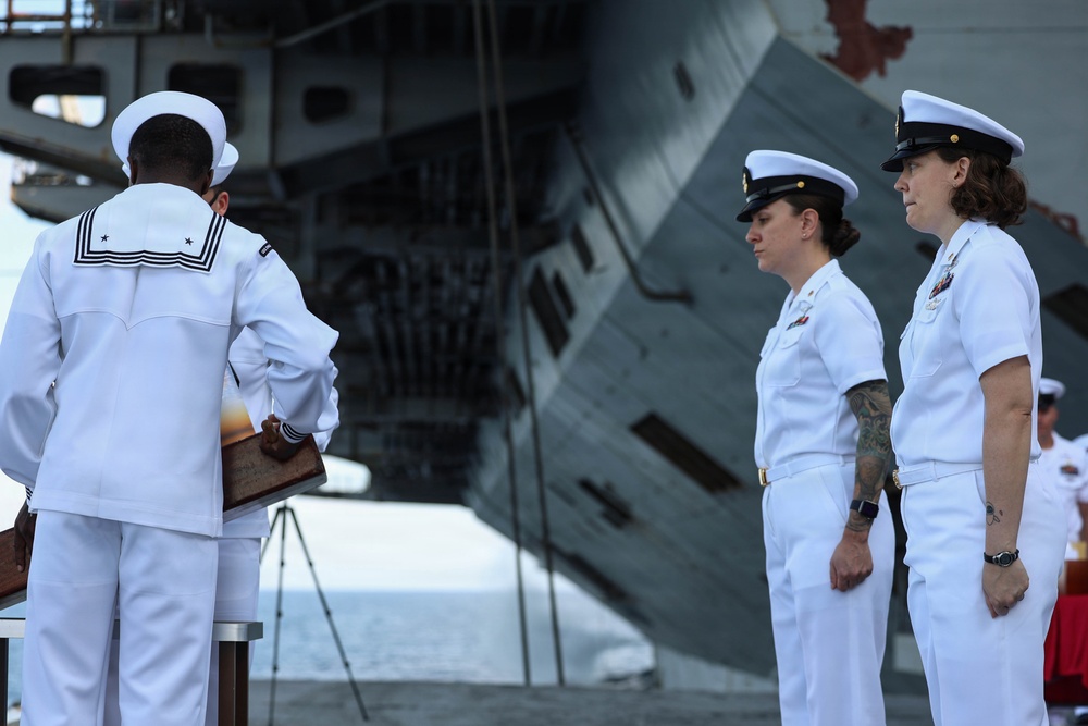 A formal U.S. Navy ceremony is taking place outdoors in front of a large naval vessel, likely an aircraft carrier. Three naval personnel in white dress uniforms are visible, with one enlisted sailor presenting or handling a wooden plaque or award to two female officers standing at attention. The setting and attire suggest a change of command, retirement, or commendation ceremony.