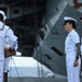 A formal U.S. Navy ceremony is taking place outdoors in front of a large naval vessel, likely an aircraft carrier. Three naval personnel in white dress uniforms are visible, with one enlisted sailor presenting or handling a wooden plaque or award to two female officers standing at attention. The setting and attire suggest a change of command, retirement, or commendation ceremony.