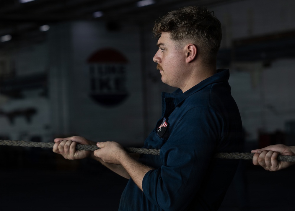 A young male in a dark navy uniform is holding a thick rope with both hands while looking to the side. A patch resembling a US flag is visible on his chest. The background contains partially visible text and a circular emblem, suggesting a military or naval vessel environment.