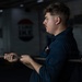 A young male in a dark navy uniform is holding a thick rope with both hands while looking to the side. A patch resembling a US flag is visible on his chest. The background contains partially visible text and a circular emblem, suggesting a military or naval vessel environment.