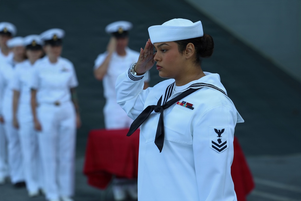 A U.S. Navy sailor in dress white uniform is rendering a salute in the foreground during what appears to be a formal military ceremony. The sailor displays a Petty Officer Third Class rating badge on the left sleeve and service ribbons on the chest. Additional sailors in dress whites are visible in the blurred background, standing in formation.