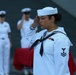 A U.S. Navy sailor in dress white uniform is rendering a salute in the foreground during what appears to be a formal military ceremony. The sailor displays a Petty Officer Third Class rating badge on the left sleeve and service ribbons on the chest. Additional sailors in dress whites are visible in the blurred background, standing in formation.