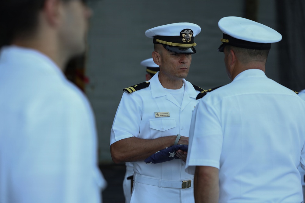 A U.S. Navy officer in white dress uniform holds a folded American flag during what appears to be a formal military ceremony. The central figure, whose name tag indicates a technical officer role, is engaged with another officer while additional personnel are visible in the background and foreground. The scene is consistent with a flag presentation or retirement ceremony.