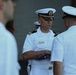 A U.S. Navy officer in white dress uniform holds a folded American flag during what appears to be a formal military ceremony. The central figure, whose name tag indicates a technical officer role, is engaged with another officer while additional personnel are visible in the background and foreground. The scene is consistent with a flag presentation or retirement ceremony.
