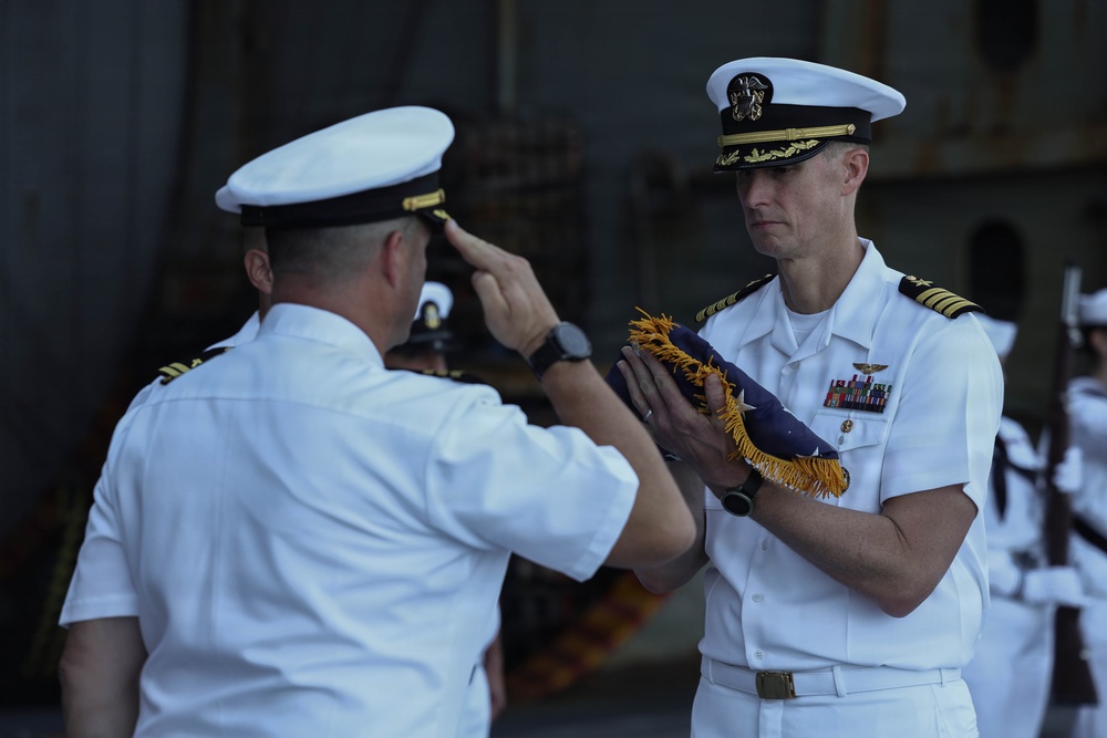 Two U.S. Navy officers in white dress uniforms are engaged in a formal military ceremony, likely a change of command or retirement ceremony. One officer is saluting while the other holds a folded flag with gold fringe, a common element in official military ceremonies. A formation of sailors in white uniforms is visible in the background.