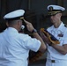 Two U.S. Navy officers in white dress uniforms are engaged in a formal military ceremony, likely a change of command or retirement ceremony. One officer is saluting while the other holds a folded flag with gold fringe, a common element in official military ceremonies. A formation of sailors in white uniforms is visible in the background.