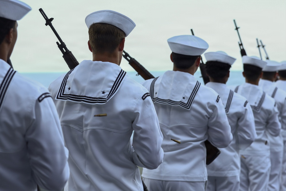 A formation of naval personnel dressed in white dress uniforms are standing in rows, each carrying rifles on their shoulders. The image appears to capture a formal military ceremony or parade, viewed from behind the sailors. The depth of field creates a receding line of uniformed figures extending into the background.