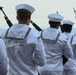 A formation of naval personnel dressed in white dress uniforms are standing in rows, each carrying rifles on their shoulders. The image appears to capture a formal military ceremony or parade, viewed from behind the sailors. The depth of field creates a receding line of uniformed figures extending into the background.