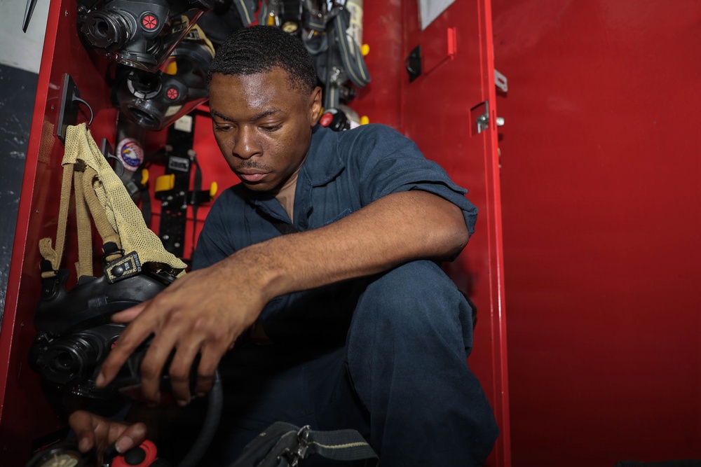 A young man in dark blue coveralls is crouched down inspecting or handling equipment inside a red storage locker or cabinet. The locker contains multiple breathing apparatus units, masks, and associated gear mounted on the interior walls. The setting appears to be a military vessel or emergency services facility based on the equipment visible.