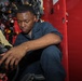 A young man in dark blue coveralls is crouched down inspecting or handling equipment inside a red storage locker or cabinet. The locker contains multiple breathing apparatus units, masks, and associated gear mounted on the interior walls. The setting appears to be a military vessel or emergency services facility based on the equipment visible.