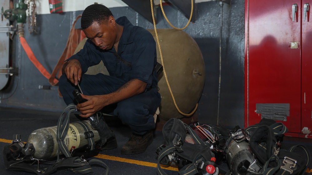A person in navy blue coveralls is crouched on the deck of what appears to be a military vessel, inspecting or maintaining breathing apparatus equipment. Multiple SCBA (Self-Contained Breathing Apparatus) units with Scott-brand air cylinders are laid out on the floor around them. The setting appears to be an interior deck area with red storage lockers and firefighting equipment visible in the background.