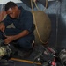 A person in navy blue coveralls is crouched on the deck of what appears to be a military vessel, inspecting or maintaining breathing apparatus equipment. Multiple SCBA (Self-Contained Breathing Apparatus) units with Scott-brand air cylinders are laid out on the floor around them. The setting appears to be an interior deck area with red storage lockers and firefighting equipment visible in the background.