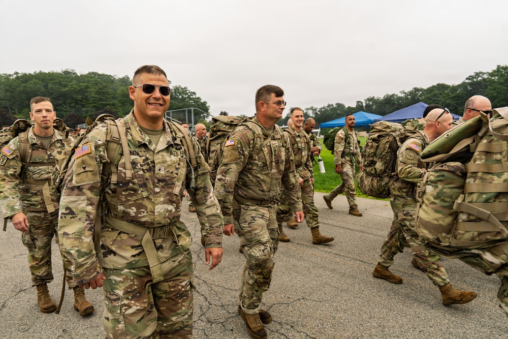 A group of U.S. Army soldiers in Operational Camouflage Pattern (OCP) uniforms are marching or walking across a paved area, carrying large rucksacks. Several soldiers in the foreground are smiling, suggesting a positive or celebratory atmosphere, possibly following a ruck march or training exercise. The setting appears to be a military installation or training ground with green grass, trees, and a blue canopy tent visible in the background.