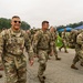 A group of U.S. Army soldiers in Operational Camouflage Pattern (OCP) uniforms are marching or walking across a paved area, carrying large rucksacks. Several soldiers in the foreground are smiling, suggesting a positive or celebratory atmosphere, possibly following a ruck march or training exercise. The setting appears to be a military installation or training ground with green grass, trees, and a blue canopy tent visible in the background.