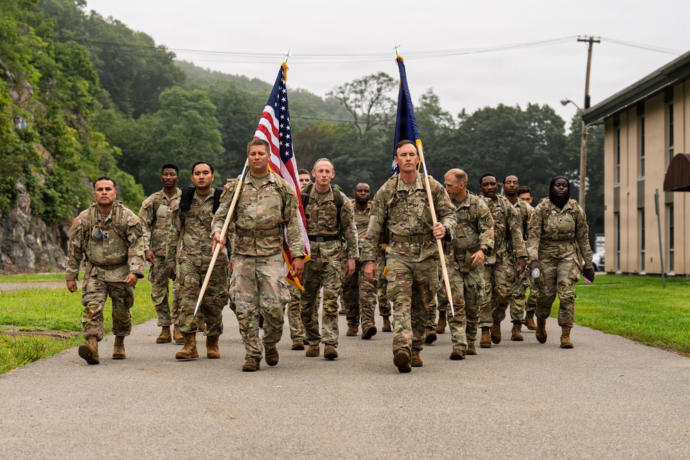 A group of U.S. military personnel in MultiCam/OCP camouflage uniforms march in formation along a paved road. Two soldiers at the front carry flags — the U.S. national flag and what appears to be a unit or service flag. The setting appears to be a military installation surrounded by wooded hills under overcast skies.