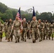 A group of U.S. military personnel in MultiCam/OCP camouflage uniforms march in formation along a paved road. Two soldiers at the front carry flags — the U.S. national flag and what appears to be a unit or service flag. The setting appears to be a military installation surrounded by wooded hills under overcast skies.
