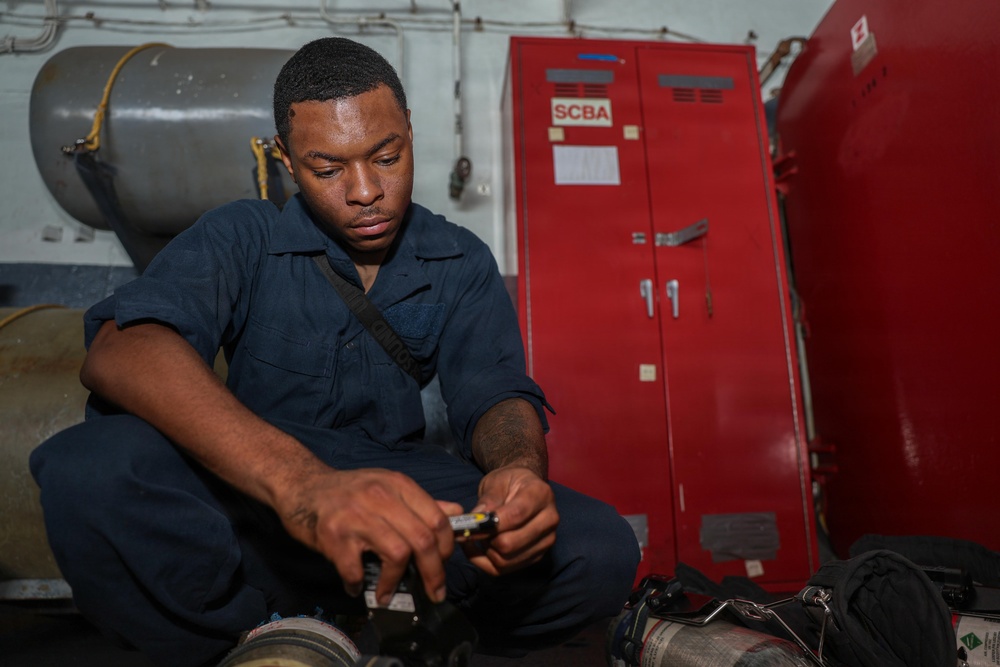 A young man in navy blue coveralls is seated on the floor, focused on handling a small piece of equipment or tool. Behind him is a red cabinet labeled 'SCBA' (Self-Contained Breathing Apparatus), indicating a military or emergency services environment, likely aboard a naval vessel. The setting suggests routine maintenance or inspection of firefighting or safety equipment.