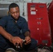 A young man in navy blue coveralls is seated on the floor, focused on handling a small piece of equipment or tool. Behind him is a red cabinet labeled 'SCBA' (Self-Contained Breathing Apparatus), indicating a military or emergency services environment, likely aboard a naval vessel. The setting suggests routine maintenance or inspection of firefighting or safety equipment.