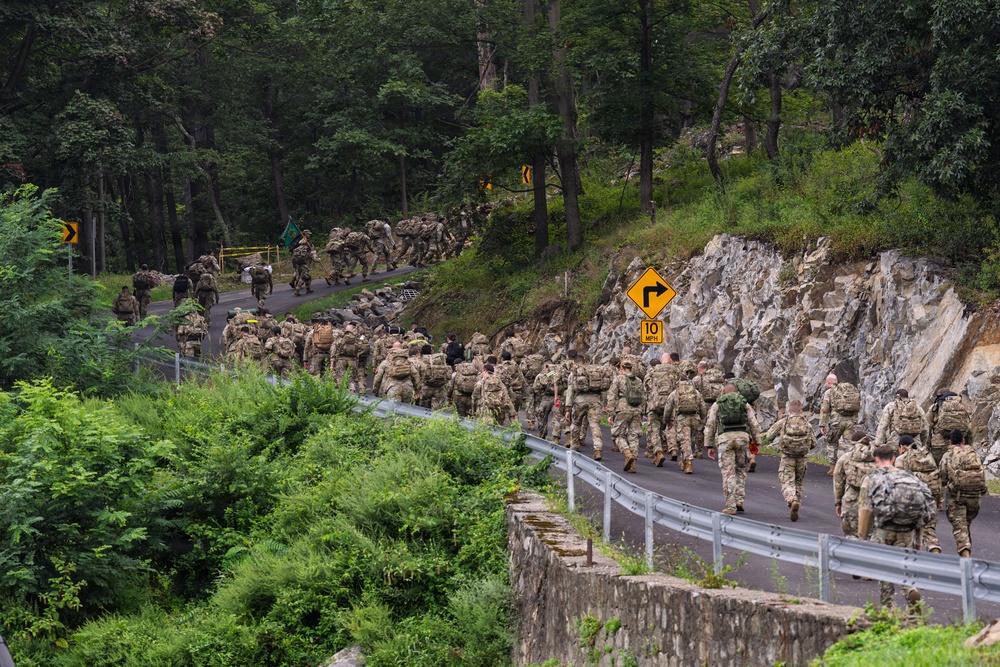 A large column of military personnel in camouflage uniforms and full gear are conducting a road march along a winding, wooded road. The group stretches far into the distance around a curve, suggesting a significant formation exercise or ruck march event. The setting appears to be a forested area with rocky outcroppings and guardrails along the roadway.