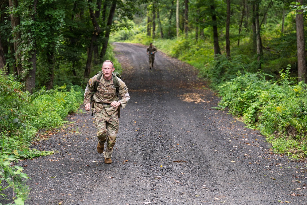 Two military personnel in camouflage uniforms are running along a narrow gravel road through a dense, green forested area. The foreground soldier is clearly visible, wearing full combat gear including a backpack, while a second soldier is visible further down the road in the background. The setting and attire suggest a military physical training exercise or ruck march.