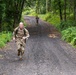 Two military personnel in camouflage uniforms are running along a narrow gravel road through a dense, green forested area. The foreground soldier is clearly visible, wearing full combat gear including a backpack, while a second soldier is visible further down the road in the background. The setting and attire suggest a military physical training exercise or ruck march.