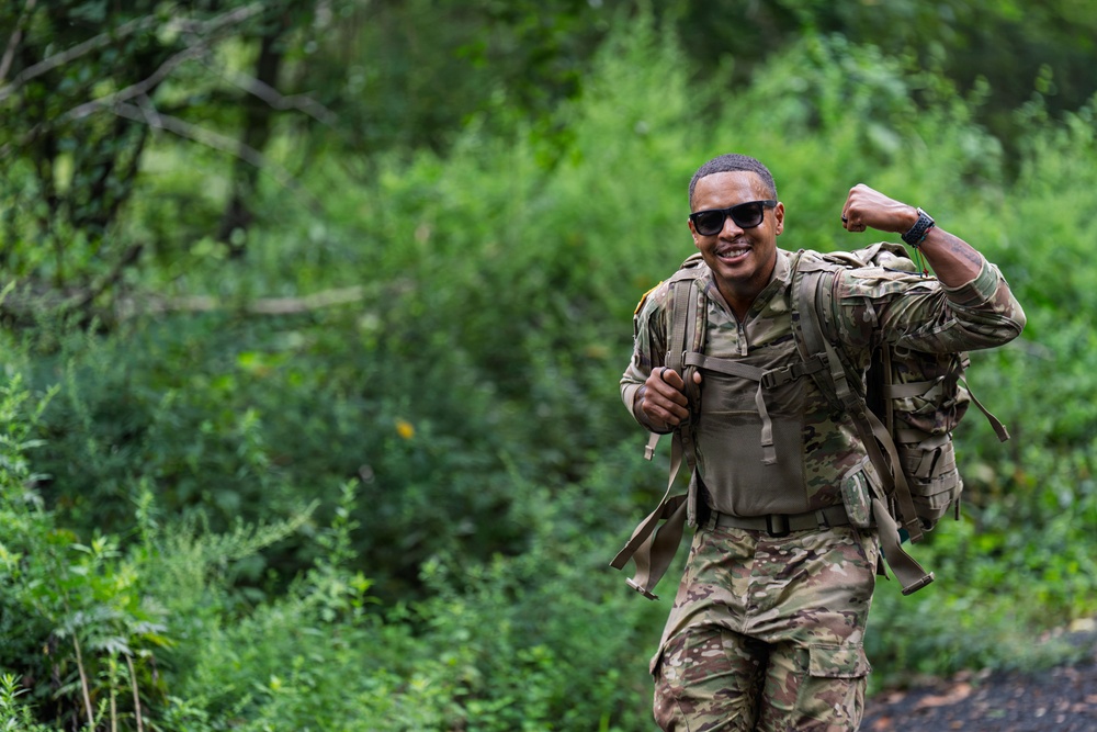 A soldier in full camouflage uniform and tactical gear is walking along a wooded trail while flexing one arm and smiling confidently. The individual is wearing a military rucksack and appears to be completing a ruck march or field training exercise. The mood is upbeat and energetic, suggesting completion of or progress through a physical fitness challenge.