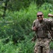 A soldier in full camouflage uniform and tactical gear is walking along a wooded trail while flexing one arm and smiling confidently. The individual is wearing a military rucksack and appears to be completing a ruck march or field training exercise. The mood is upbeat and energetic, suggesting completion of or progress through a physical fitness challenge.