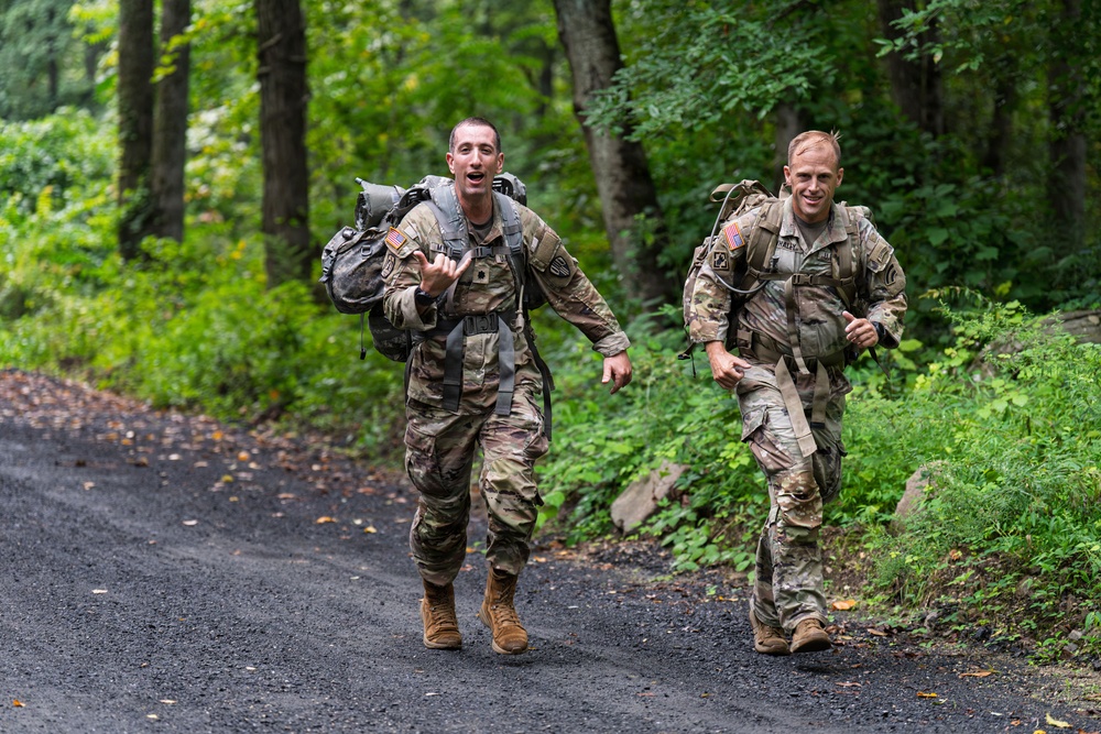 Two U.S. Army soldiers in full camouflage uniforms and tactical gear are conducting a ruck march along a paved forest road. Both soldiers are carrying large military rucksacks and appear to be moving at a brisk pace, with the lead soldier appearing to vocalize or call out. The setting is a wooded area with lush green vegetation, suggesting a training exercise environment.