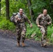 Two U.S. Army soldiers in full camouflage uniforms and tactical gear are conducting a ruck march along a paved forest road. Both soldiers are carrying large military rucksacks and appear to be moving at a brisk pace, with the lead soldier appearing to vocalize or call out. The setting is a wooded area with lush green vegetation, suggesting a training exercise environment.