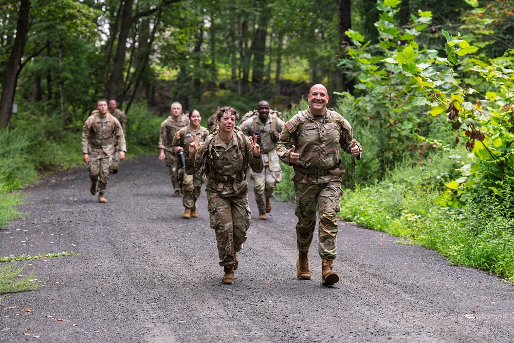 A group of military personnel in camouflage uniforms and tactical gear are conducting a ruck march or physical training exercise along a paved road surrounded by dense green forest. The two lead soldiers are visibly smiling and making positive gestures — one giving thumbs up and the other making peace signs — suggesting high morale. Several more soldiers follow behind in a loose formation.