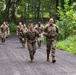 A group of military personnel in camouflage uniforms and tactical gear are conducting a ruck march or physical training exercise along a paved road surrounded by dense green forest. The two lead soldiers are visibly smiling and making positive gestures — one giving thumbs up and the other making peace signs — suggesting high morale. Several more soldiers follow behind in a loose formation.