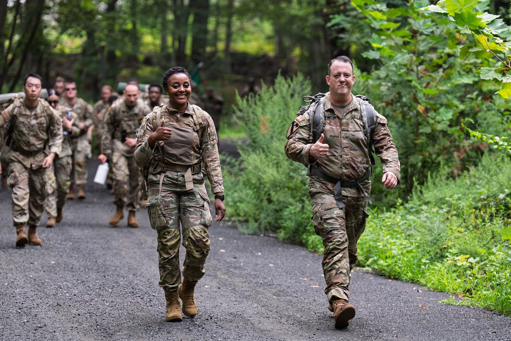A group of military personnel in camouflage uniforms are conducting a ruck march along a wooded path. Two soldiers in the foreground — one female and one male — are smiling and giving thumbs-up gestures, suggesting positive morale. A column of additional soldiers follows behind them on the same trail.