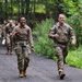 A group of military personnel in camouflage uniforms are conducting a ruck march along a wooded path. Two soldiers in the foreground — one female and one male — are smiling and giving thumbs-up gestures, suggesting positive morale. A column of additional soldiers follows behind them on the same trail.