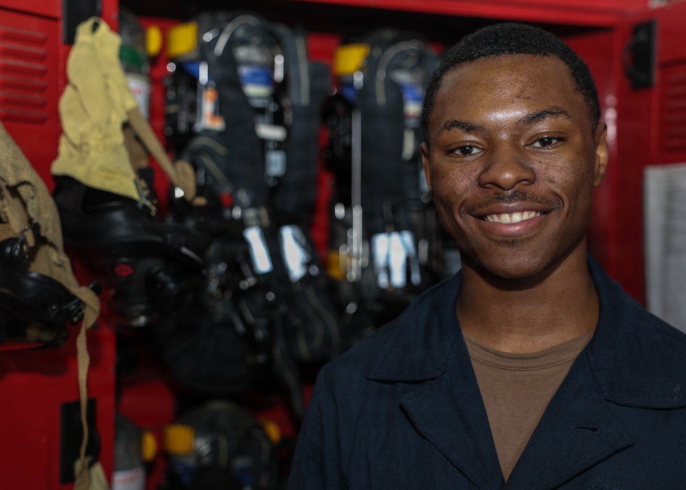 A young man in a dark navy uniform smiles confidently at the camera while standing in front of red equipment lockers. The background contains firefighting or emergency response gear, including SCBA (Self-Contained Breathing Apparatus) units and protective equipment. The setting appears to be a fire station or emergency services facility.