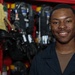 A young man in a dark navy uniform smiles confidently at the camera while standing in front of red equipment lockers. The background contains firefighting or emergency response gear, including SCBA (Self-Contained Breathing Apparatus) units and protective equipment. The setting appears to be a fire station or emergency services facility.