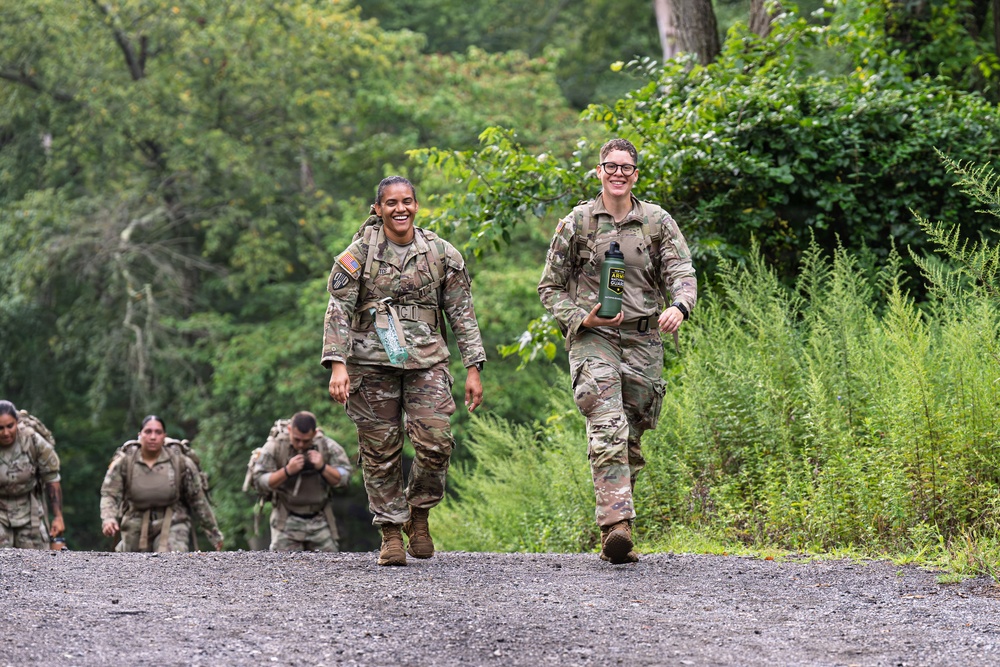A group of military personnel in U.S. Army OCP (Operational Camouflage Pattern) uniforms are conducting a ruck march along a gravel path surrounded by lush green vegetation. Two female soldiers lead the formation in the foreground, both smiling and carrying water bottles, while additional soldiers follow behind. The setting appears to be a wooded training area during warm weather conditions.