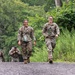 A group of military personnel in U.S. Army OCP (Operational Camouflage Pattern) uniforms are conducting a ruck march along a gravel path surrounded by lush green vegetation. Two female soldiers lead the formation in the foreground, both smiling and carrying water bottles, while additional soldiers follow behind. The setting appears to be a wooded training area during warm weather conditions.