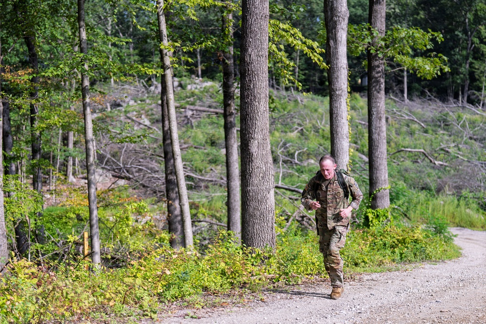 A soldier in military camouflage uniform is running along a gravel path through a forested area. The individual is carrying a backpack and appears to be engaged in a physical training or ruck march exercise. The background shows a mix of standing trees and fallen/cleared timber on a rocky hillside.