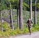 A soldier in military camouflage uniform is running along a gravel path through a forested area. The individual is carrying a backpack and appears to be engaged in a physical training or ruck march exercise. The background shows a mix of standing trees and fallen/cleared timber on a rocky hillside.