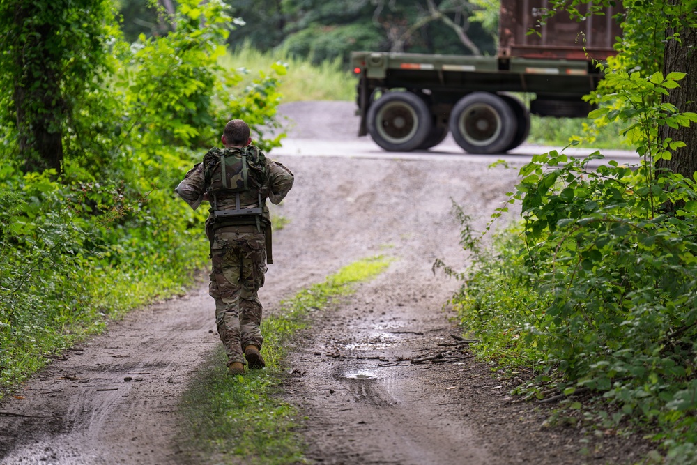 A soldier in camouflage uniform and tactical gear walks away from the camera down a muddy dirt road flanked by dense green vegetation. In the background, a military-style truck is visible on a paved road at the end of the dirt path. The scene appears to be a training or operational environment in a temperate, wooded area.