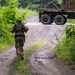 A soldier in camouflage uniform and tactical gear walks away from the camera down a muddy dirt road flanked by dense green vegetation. In the background, a military-style truck is visible on a paved road at the end of the dirt path. The scene appears to be a training or operational environment in a temperate, wooded area.