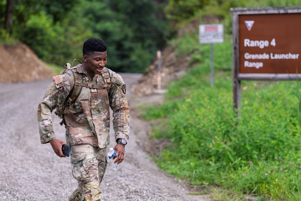 A U.S. Army soldier in OCP (Operational Camouflage Pattern) uniform walks along a gravel road near Range 4, a Grenade Launcher Range at a military training installation. The soldier is carrying a water bottle and a phone while wearing a tactical backpack, appearing relaxed and smiling. The setting is a lush, green outdoor military training area.