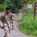 A U.S. Army soldier in OCP (Operational Camouflage Pattern) uniform walks along a gravel road near Range 4, a Grenade Launcher Range at a military training installation. The soldier is carrying a water bottle and a phone while wearing a tactical backpack, appearing relaxed and smiling. The setting is a lush, green outdoor military training area.
